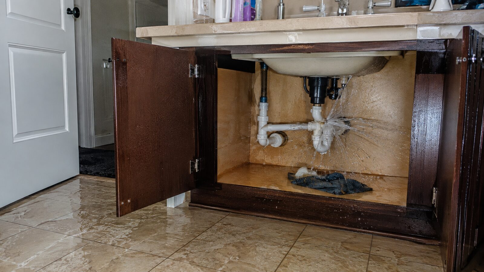 Water spraying from a split supply line inside a bathroom vanity cabinet, soaking the cabinet base and running out across the tile floor